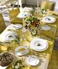 A beautifully set dining table with a yellow tablecloth, white plates, cutlery, glassware, candles, and a centerpiece of vegetables and herbs. Produktbild-Vorschau