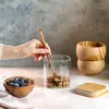 A hand is shown stirring granola in a glass jar with a wooden spoon, next to a bowl of granola and blueberries. Wooden bowls and a bamboo lid are also visible. Náhled obrázku produktu