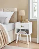 A white nightstand with a drawer and open shelf sits next to a bed. Produktbild-Vorschau