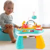 A baby is sitting on a potty chair next to a colorful interactive music table. The baby is looking at the camera and smiling. The music table has various buttons, keys, and toys on it. Produktbild-Vorschau