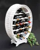 A white wooden wine rack shaped like a barrel, filled with wine bottles, stands against a black background. A green plant is to the right of the wine rack. Produktbild-Vorschau