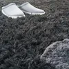 A close-up view of a pair of gray slippers resting on a dark gray, shaggy rug. Produktbild-Vorschau