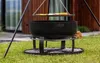 A large black fire pit with a grill rack above it, set up on a grassy lawn in front of a modern house. Produktbild-Vorschau