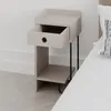 A modern beige nightstand with an open drawer, black legs, and a shelf, placed next to a bed on a light wood floor. Produktbild-Vorschau