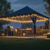 An outdoor patio scene at dusk, featuring a dark blue gazebo decorated with string lights, a dining table set for a meal, and a grill. Produktbild-Vorschau