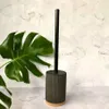 A toilet brush with a black handle and a ribbed gray holder with a wooden base, placed in front of a hexagonal marble-patterned tile wall. Anteprima dell’immagine prodotto