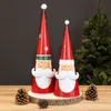 Two Santa Claus decorative figures are displayed on a wooden stand, with pine cones in the foreground and a Christmas tree in the background. Produktbild-Vorschau