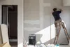 A construction worker is installing drywall on a wall while standing on a ladder. A portable heater is on the floor nearby. Produktbild-Vorschau
