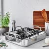 A stainless steel roasting pan with a rack is displayed on a kitchen counter, with a potted herb, a pepper grinder, a cutting board, and kitchen utensils in the background. Produktbild-Vorschau