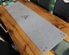 A gray table runner with star cutouts on the ends and a black graphic design in the center is laid on a wooden dining table. Produktbild-Vorschau
