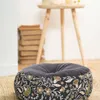 A round floor cushion with a floral pattern sits on a woven rug in front of a stack of books and a wooden side table. Produktbild-Vorschau
