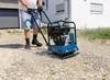 A person is using a blue plate compactor on a gravel surface in front of a house. Náhľad obrázku produktu