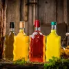 Four decorative glass bottles filled with colorful liquids are arranged on a rustic wooden surface, with moss and straw scattered around. The background is a weathered wooden wall. Produktbild-Vorschau