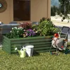 A raised garden bed filled with various plants and flowers, situated on a grassy area next to a patio with outdoor furniture. Gardening tools like a watering can and a hose reel are also visible. Voir les images du produit