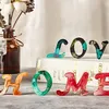Resin letters spelling 'HOME' and 'LOVE' are displayed on a surface, with a vase of flowers in the background. Produktbild-Vorschau