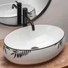 A white oval sink with black leaf pattern and a black faucet, placed on a wooden surface. Náhled obrázku produktu