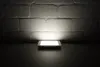 A square LED panel light is placed on a dark surface, illuminating a white brick wall. Produktbild-Vorschau