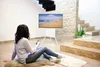 A woman sits on the floor in front of a TV stand displaying a beach scene, with stairs and a stone wall in the background. Voir les images du produit
