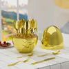 A gold cutlery set is displayed on a checkered tablecloth, with a blurred background of a window view. Produktbild-Vorschau