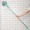 A person's arm is holding a long-handled broom with a fan-shaped brush head, angled towards a white tiled wall. Náhľad obrázku produktu