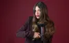 A woman holds a hairdryer in front of her hair against a red background. Náhled obrázku produktu