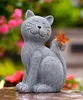 A gray cat figurine with a butterfly on its tail sits outdoors with blurred pink flowers in the background. Produktbild-Vorschau