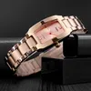 A rose gold women's watch with a pink dial is displayed on a dark, textured surface, partially resting on a black block. Produktbild-Vorschau