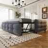 A modern bedroom with a gray upholstered sofa bed partially extended, revealing its metal frame. The room features large windows with vertical blinds, a chandelier, framed artwork on the wall, and a patterned rug on a wooden floor. Produktbild-Vorschau