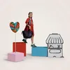 A young girl in a red dress walks up steps made of colorful blocks, holding a black quilted bag. A large lollipop is on a stick to her left, and a drawing of a market stall is to her right. Produktbild-Vorschau