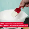 A hand is holding a red scoop with flour over a white bowl. Náhľad obrázku produktu