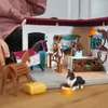 A child's hands are visible in the upper left corner, interacting with a toy horse stable playset. The playset includes toy figures of people, a horse, a dog, and various accessories like a hay bale and a saddle stand. The scene depicts a miniature horse s Produktbild-Vorschau
