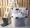 Two transparent storage boxes are stacked, with a ladder and wooden pallets in the background. A pair of grey boots are in the foreground. Produktbild-Vorschau