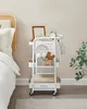 A white rolling cart filled with various items, placed next to a bed with a rattan headboard. Náhľad obrázku produktu