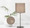 A table lamp with a taupe fabric shade and a silver metal base is shown on a white surface next to a white decorative vase with a plant. Voir les images du produit