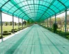 A long, covered walkway with a green translucent roof and a green path, lined with trees and manicured bushes. Produktbild-Vorschau