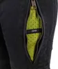 A close-up view of a zipper on black pants, revealing a bright green mesh lining with a black tag that has white text. Produktbild-Vorschau
