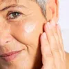 A close-up shot of a woman's face as she applies a cream to her cheek. Anteprima dell’immagine prodotto