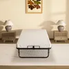 A foldable bed with a white mattress is placed in the middle of a room, flanked by two nightstands with lamps, and a painting hangs on the wall above. Voir les images du produit