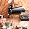 A hand pours coffee from a black and silver coffee pot into a decorative mug on a wooden table. Náhled obrázku produktu