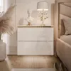 A bedroom scene featuring a white high-gloss nightstand with gold accents, a decorative bust, a lamp, and a vase with dried flowers. Produktbild-Vorschau