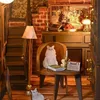 A miniature dollhouse scene depicting a cozy room with stairs, a fireplace, furniture, and decorative items, including two cat figurines. Voir les images du produit
