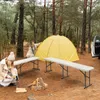 Two people are camping in a forest with a yellow tent, two benches, and a campfire. Produktbild-Vorschau