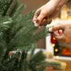 Hands adjusting the branches of an artificial Christmas tree with a fireplace in the background. Produktbild-Vorschau