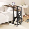 A C-shaped side table with a charging station and storage pockets is placed next to a white sofa, with a small round white table and a plush toy in the background. Produktbild-Vorschau