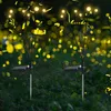 Two solar-powered firefly lights are planted in the ground at night, with blurred yellow lights in the background. Produktbild-Vorschau
