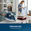 A woman is ironing clothes in a well-lit room with shelves of clothes and a sofa in the background. An iron is on an ironing board in the foreground. Náhľad obrázku produktu