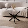 A clear glass coffee table with a black base is placed on a wooden floor in front of a curved, light-colored sofa. A book, a decorative sphere, and a vase with dried flowers are on the sofa. Produktbild-Vorschau