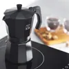 A black moka pot on a stovetop, with two cups of coffee and sugar cubes in the background. Produktbild-Vorschau