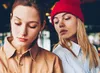 Two young women looking down at something out of frame, one wearing a red beanie. Náhľad obrázku produktu