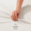 A hand is shown touching a white textured fabric, likely a bedsheet or towel, with text indicating material composition. Produktbild-Vorschau